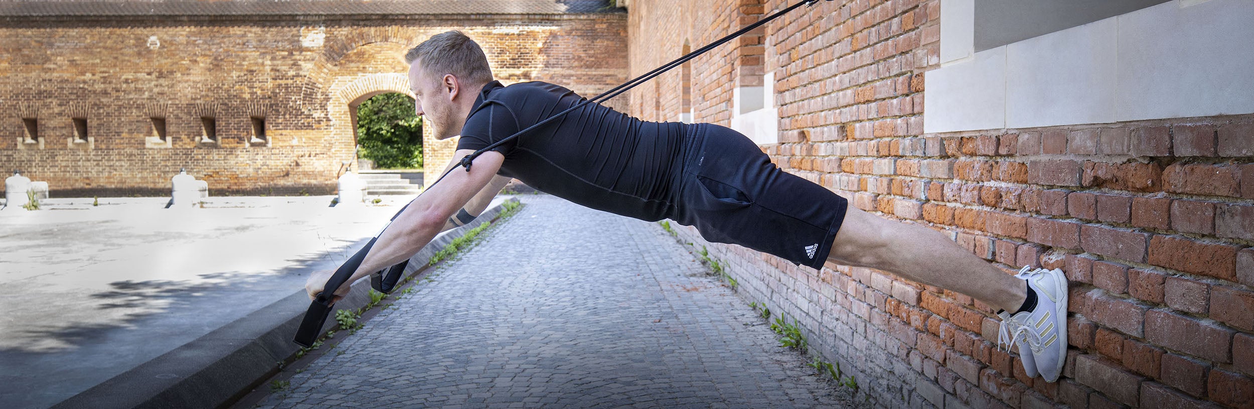 Person hängt horizontal an einem Sling-Trainer, die Füße sind an einer Hauswand abgestützt