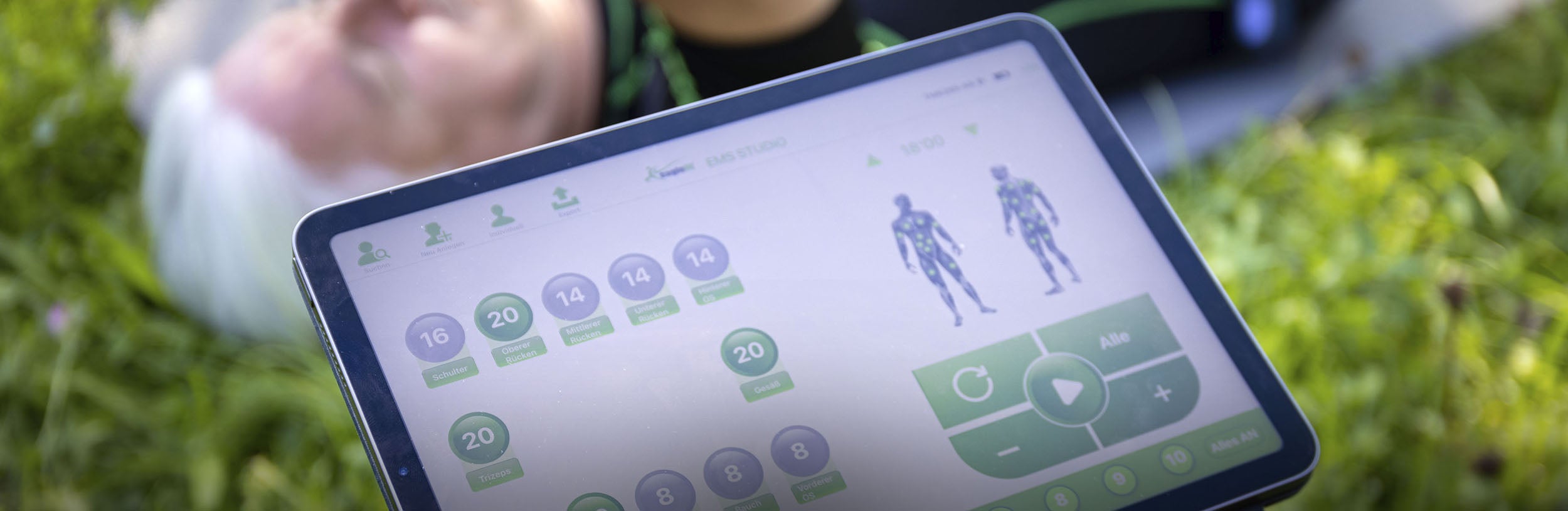 Blick auf ein Display mit der Trainings App von eaglefit. Im Hintergrund sieht man eine Person liegend, die eine Übung durchführt.