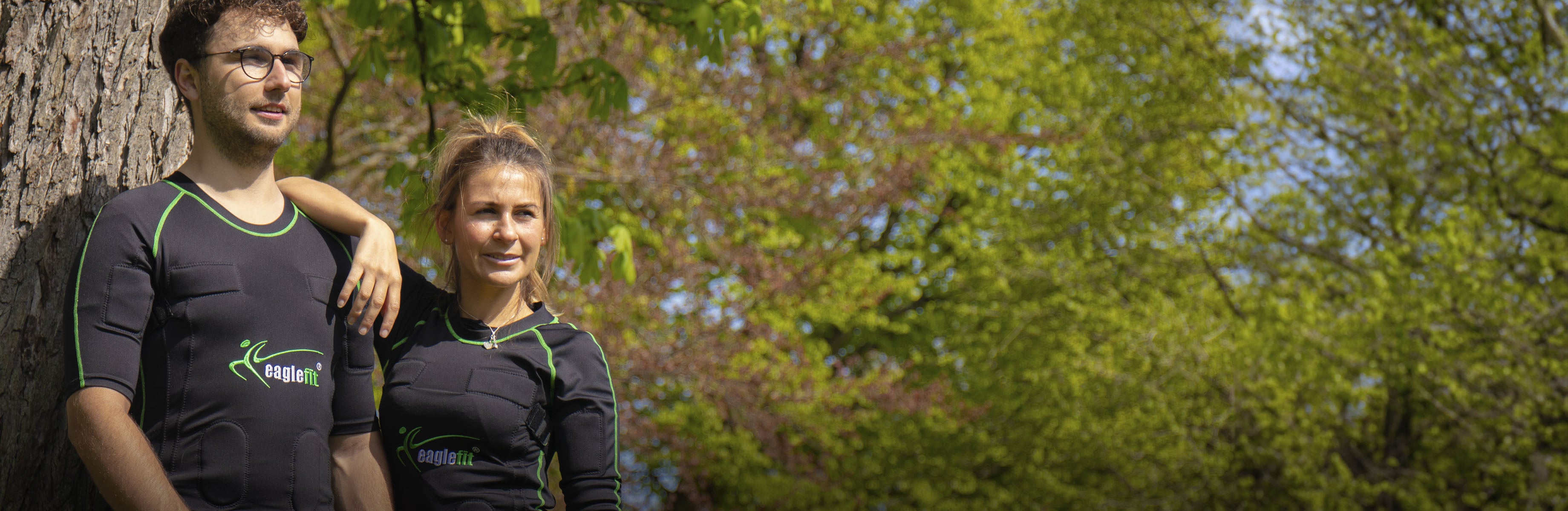 Zwei Personen in EMS-Anzügen lehnen an einem Baum und blicken rechts am Betrachter vorbei
