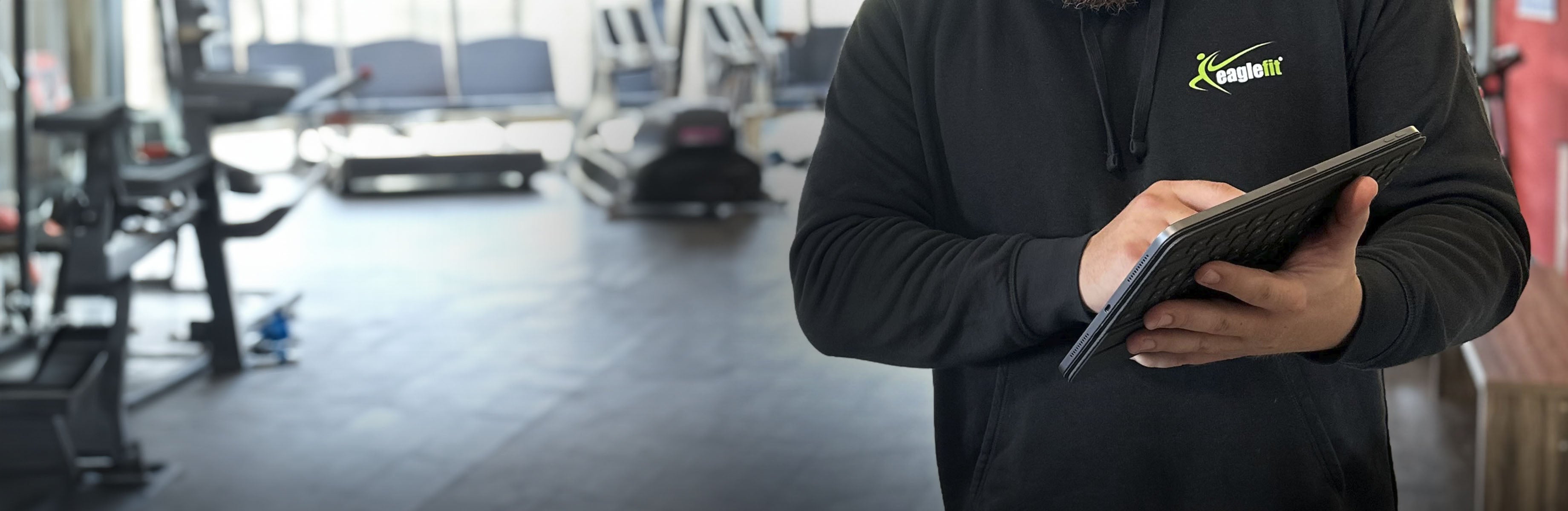 Person mit Tablet im eaglefit Shirt und im Hintergrund eine Fitness-Studio-Situation